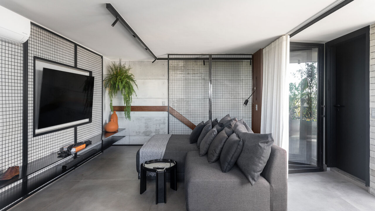 Dúplex industrial de 290 m² une piscina de vidro e área para ver jogos. Projeto Morada 31.12. Na foto, sala de TV com estrutura metálica na parede e sofá cinza.