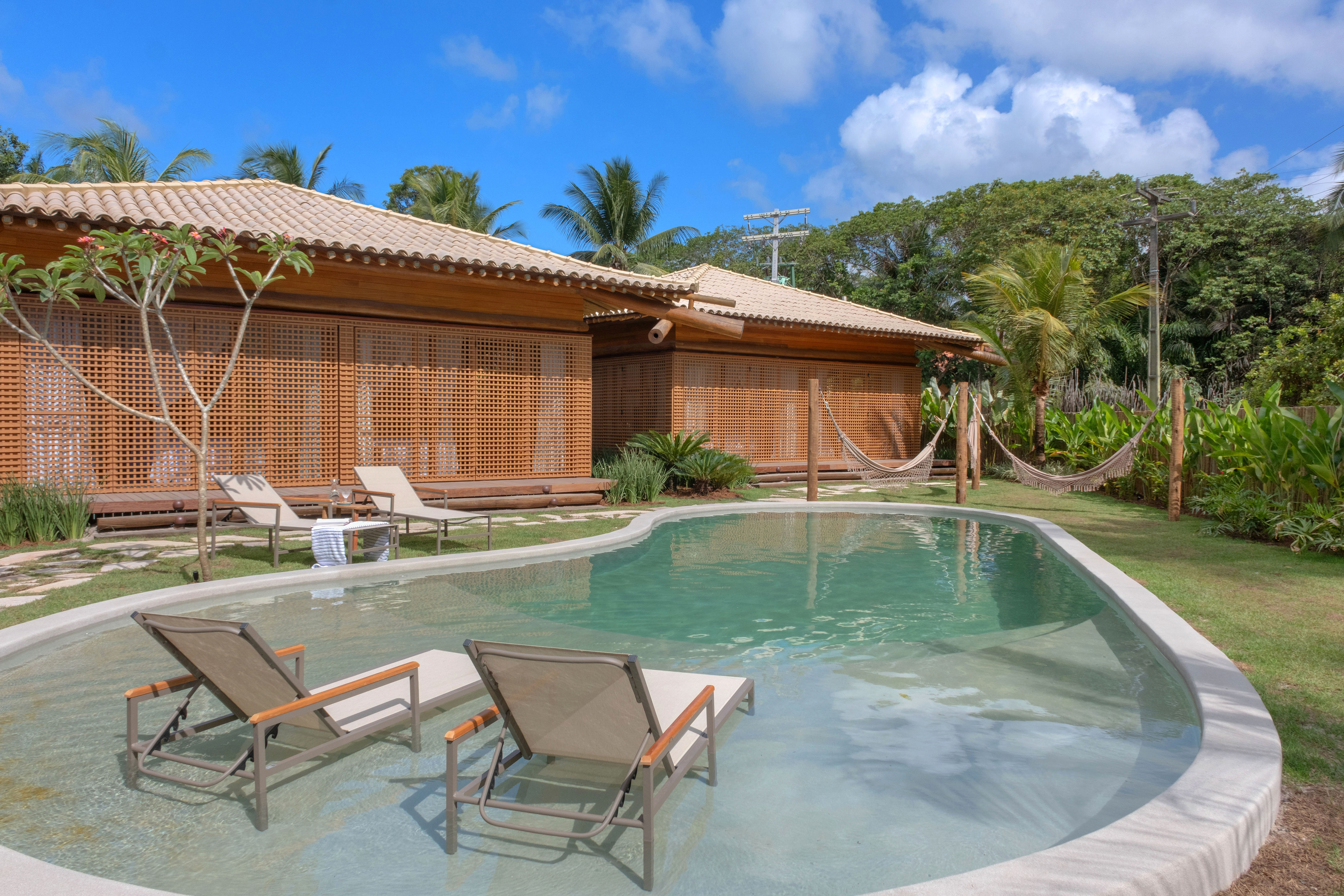 Casa de praia com estrutura de madeira possui piscina orgânica e redário. Projeto de David Bastos. Na foto, jardim com piscina, redes e jardim.